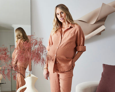 A pregnant woman in a loose, peach-colored outfit stands indoors near a modern white vase with dried pink foliage. She is reflected in a tall mirror behind her, and abstract wall art hangs nearby.