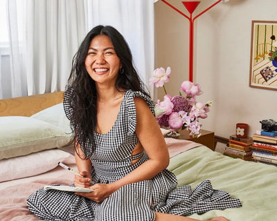 A smiling woman in a black-and-white gingham dress sits on a bed holding a notebook. Behind her are pillows, flowers in a vase, a red lamp, books, and framed artwork on the wall.