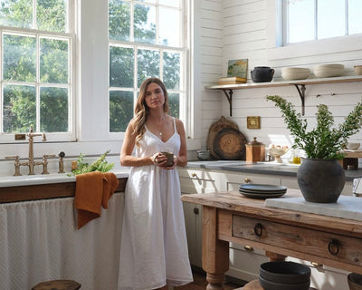 A woman in a white dress stands in a bright, rustic kitchen with large windows, wooden shelves, and a wooden table. She holds a mug and is surrounded by plants, dishes, and natural light.