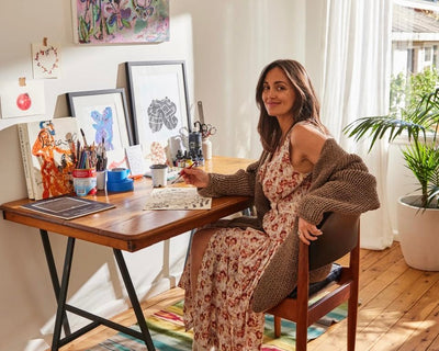 A woman in a floral dress and cardigan sits at a wooden desk with art supplies, sketches, and framed artwork. Sunlight streams through a window, and a potted plant is in the background. She smiles at the camera.