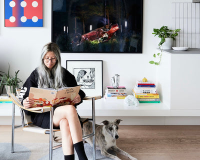 A woman with long gray hair sits in a modern chair reading a cookbook. A dog lies beside her on the floor. Behind them are books, plants, and colorful artwork on a white shelf and wall in a bright, stylish living room.