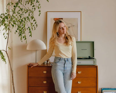 A woman with long blonde hair, wearing a yellow top and light blue jeans, stands in a cozy room by a wooden dresser with a lamp, framed art, a plant, and a green record player on top.
