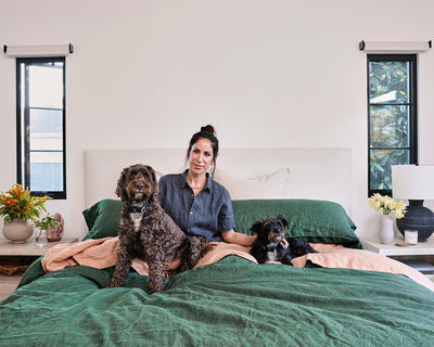 A woman sits on a bed with green covers, smiling, with a large brown dog and a small black dog beside her. There are flowers and decor on nightstands next to windows on each side of the bed.