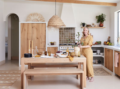 A smiling woman in a tan jumpsuit stands in a bright, modern kitchen with wood accents, setting a rustic dining table adorned with plates, glasses, and a branch in a vase. Green plants and natural light fill the space.