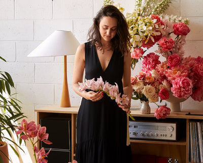 A woman in a black dress stands indoors, holding a stem of pink flowers. She is surrounded by various vibrant flowers and a record player on a wooden shelf, with a white lamp and potted plant nearby.