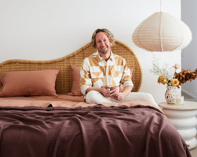 A smiling man with wavy blonde hair sits on a made bed with rust and mauve bedding, against a rattan headboard, holding a mug. A paper lantern and a vase with flowers are on a bedside table beside him.