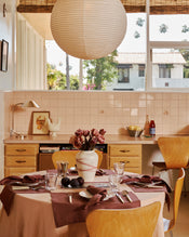 Dining room with a table set for a meal, flowers in the center, and a view of a house from the window. On the table is the 100% French flax tablecloth in terracotta and four napkins and placemats in cacao. - view