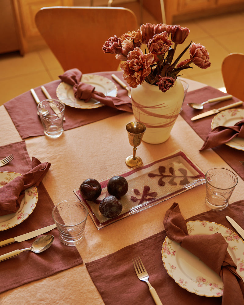 Set dining table with a terracotta linen tablecloth and cacao placemats and napkins, with floral arrangement, plates, glasses, and cutlery. 