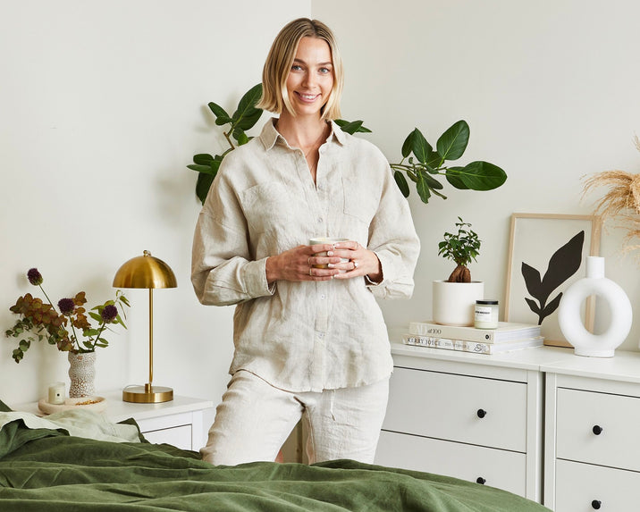 A woman in light-colored, casual clothes stands by a bed holding a cup, smiling. She is in a cozy, modern bedroom with plants, books, and minimalist decor on white dressers.