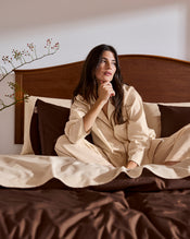 A woman wears the Bed Threads Vanilla & Espresso Piped Long Sleeve Shirt, sitting thoughtfully on a bed with brown and beige bedding and a branch with small berries in the background. - view