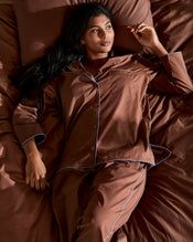 A woman with long dark hair reclines on a bed with brown sheets, wearing Bed Threads’ Espresso & Sky Piped Pants—breathable cotton pajamas with white piping and an elastic waist—looking relaxed as she gazes thoughtfully upward. - view