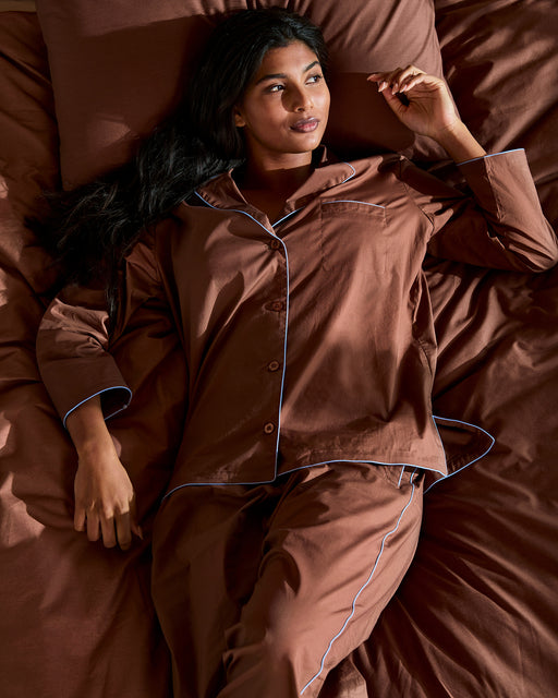 A woman with long dark hair reclines on a bed with brown sheets, wearing Bed Threads’ Espresso & Sky Piped Pants—breathable cotton pajamas with white piping and an elastic waist—looking relaxed as she gazes thoughtfully upward.