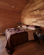 A cozy bedroom with a bed dressed in 100% french flax linen cacao duvet and pillowcases, and fitted and flat sheets in a brown-taupe colour Portobello, set against a wall and ceiling made of warm wood paneling. A small bedside table holds a glass and a small decorative object. - view