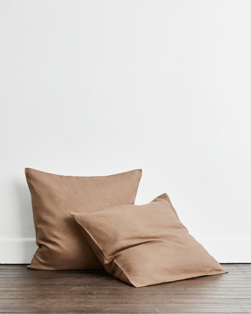 Two square european pillowcases in a warm taupe-brown 100% french flax linen fabric, one standing upright and one leaning in front, set on a dark wood floor against a plain white wall.