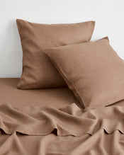 A close up image of two brown-taupe european pillowcases and sheets in 100% french flax linen, resting against a white wall.  - view