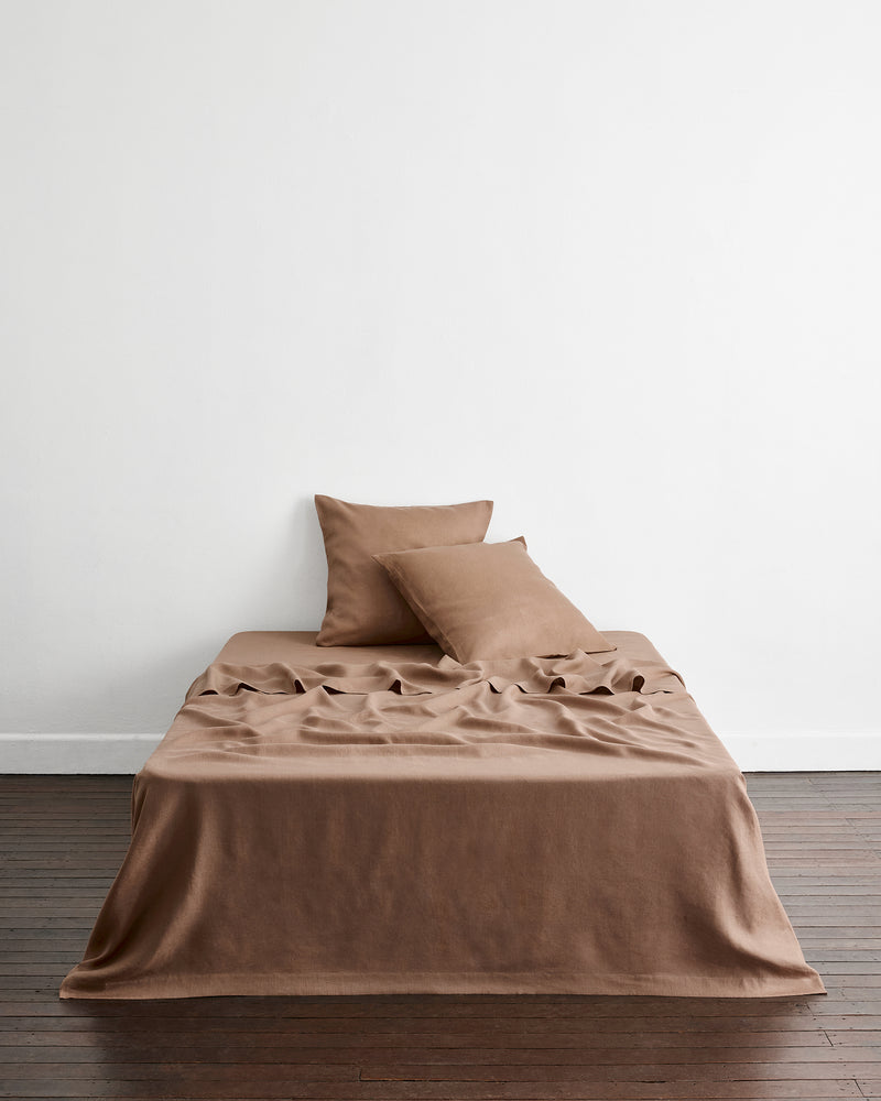 A bed sits in a room with dark wooden floors and a white wall, dressed in warm taupe-brown 100% french flax linens. A flat sheet drapes onto the floor and two european pillowcases lean against the white wall.