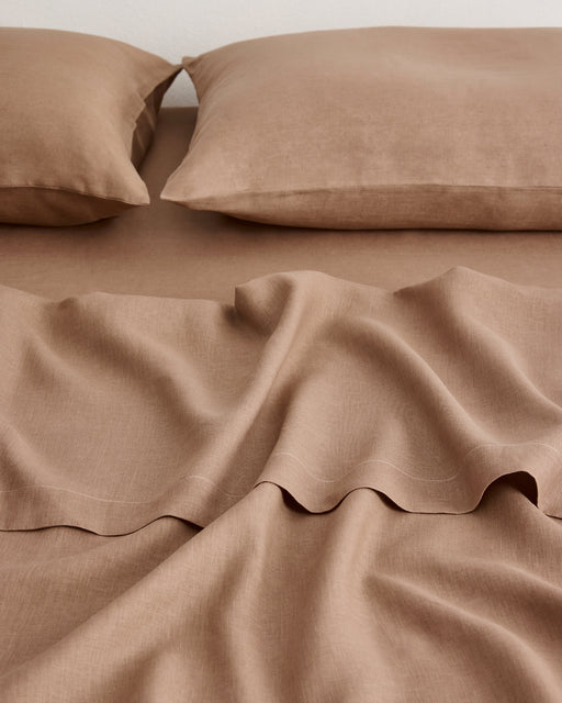 A close-up of a bed made with matching taupe-brown sheets and two pillowcases, showing soft linen fabric folds and a clean, coordinated look.