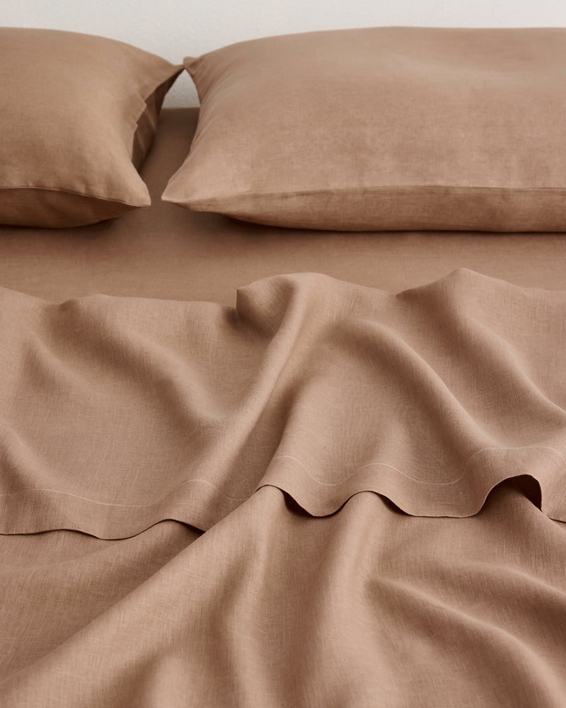 A close-up of a bed made with matching taupe-brown sheets and two pillowcases, showing soft linen fabric folds and a clean, coordinated look.