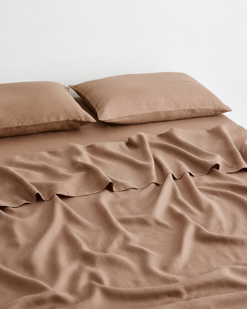 A bed dressed in a matching brown-taupe 100% french flax linen bedding set with two pillowcases and a sheet against a plain light background.