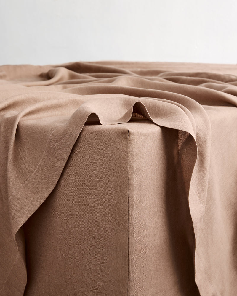 A close-up of a draped taupe-brown flat sheet in 100% french flax linen resting on the corner of a fitted sheet, showing its soft texture and folds against a neutral light background.