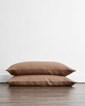 Two king size pillowcases in a brown taupe shade called Portobello available in 100% french flax linen, stacked on top of each other on a dark wood floor against a plain white wall. - view
