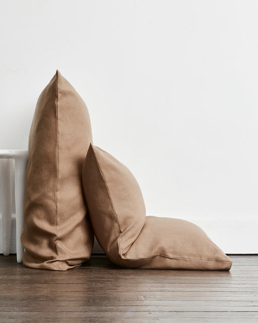 Two brown-taupe standard pillows in 100% french flax linen, one standing upright and one leaning against it, placed on a dark wood floor against a white wall.