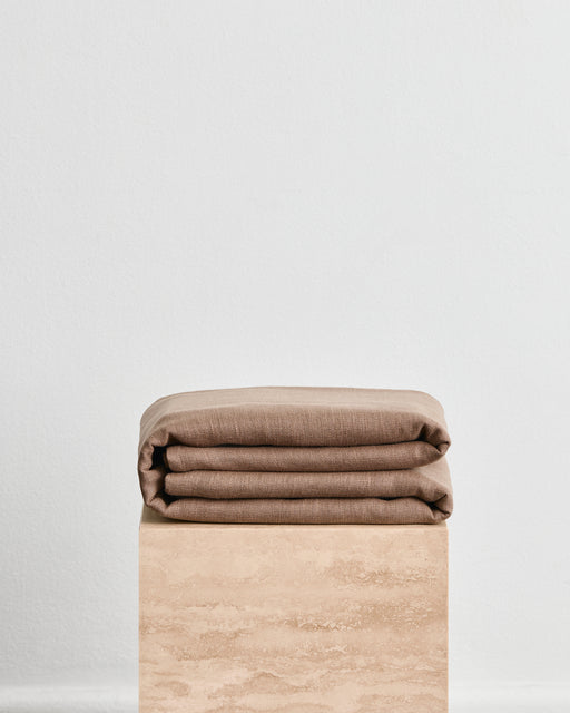 A folded warm brown throw in 100% french flax linen, folded neatly on a light beige stone pedestal against a plain white wall.