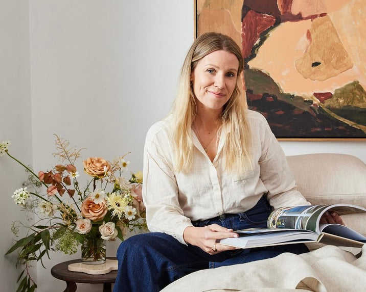 A woman with long blonde hair, wearing a light shirt and jeans, sits on a beige couch holding an open magazine. Behind her is a vase of flowers on a small table and an abstract painting on the wall.