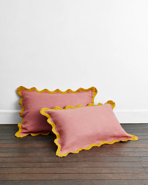 Two Pink Clay & Turmeric 100% French Flax Linen Scalloped Pillowcases by Bed Threads are displayed on a wooden floor against a plain white wall.