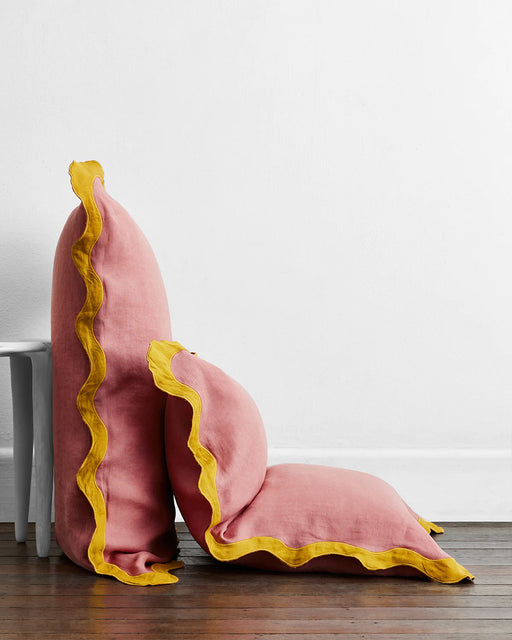 Two Pink Clay & Turmeric 100% French Flax Linen Scalloped Pillowcases by Bed Threads, featuring yellow ruffled edges, rest against a white wall—one upright on a stool, the other flat—bringing Jean Royère-inspired charm to the room.
