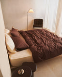 A bed styled with the Bed Threads Espresso & Vanilla Organic Cotton Bedding Bundle shows cream bedding and dark brown accents. Pillowcases and a matching percale duvet cover rest atop, while a bedside table holds a vase next to a round table in view.