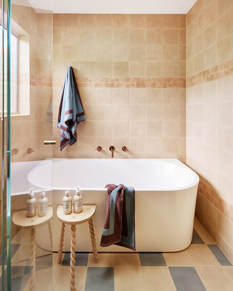 Modern bathroom with tan tiles, a white soaking tub, and gold fixtures. Two stools hold bottles, while the Cacao & Lagoon Two-Toned 100% Cotton Terry Bath Mat by Bed Threads adds comfort. Muted towels hang nearby as sunlight brightens the space.
