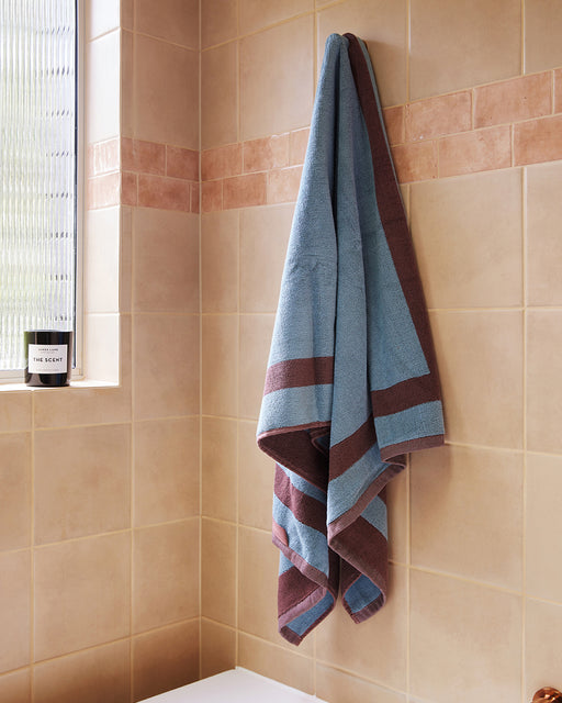 The Cacao & Lagoon Two-Toned 100% Cotton Terry Bath Towel by Bed Threads hangs on a hook against beige tiles, near a frosted window and a candle on the windowsill.