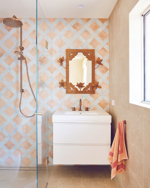 A bathroom with beige and blue checkered tile walls, a wooden framed mirror above a white sink, a bronze faucet, a glass shower with rain showerhead, and a Bed Threads Terracotta & Pink Clay Two-Toned 100% Cotton Terry Hand Towel on a hook.