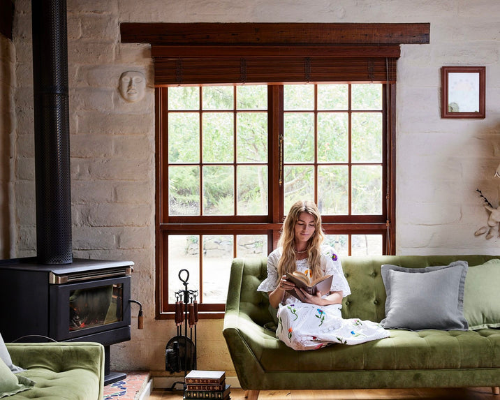 A woman sits on a green sofa in a cozy living room, reading a book near a large window with wooden blinds. A wood-burning stove and fireplace tools are beside her, with sunlight streaming in from outside.