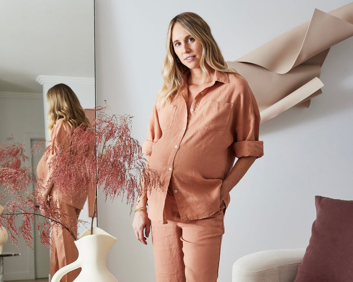 A pregnant woman in a loose, peach-colored outfit stands indoors near a modern white vase with dried pink foliage. She is reflected in a tall mirror behind her, and abstract wall art hangs nearby.