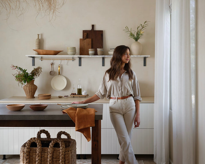 A woman stands in a bright, minimalist kitchen with neutral decor, looking out a window. The kitchen features open shelving, wooden accents, ceramics, and woven baskets for storage.