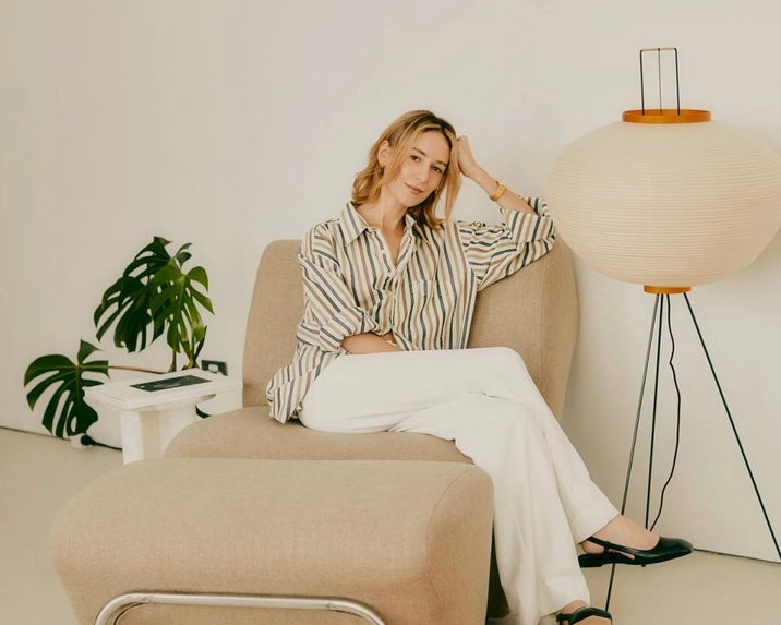 A woman with light hair sits on a beige chair, wearing a striped shirt and white pants. She is resting her head on her hand and smiling. A round paper floor lamp and a small white table with a plant are nearby.