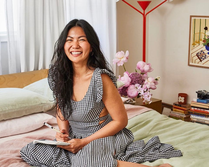 A smiling woman in a black-and-white gingham dress sits on a bed holding a notebook. Behind her are pillows, flowers in a vase, a red lamp, books, and framed artwork on the wall.