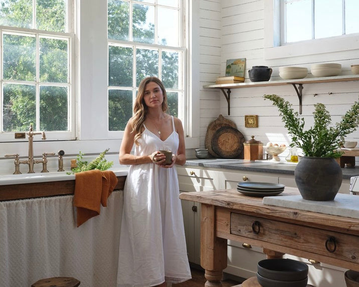 A woman in a white dress stands in a bright, rustic kitchen with large windows, wooden shelves, and a wooden table. She holds a mug and is surrounded by plants, dishes, and natural light.