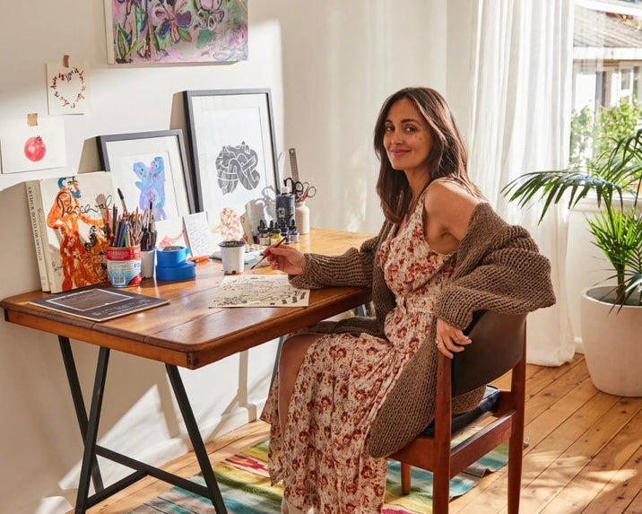 A woman in a floral dress and cardigan sits at a wooden desk with art supplies, sketches, and framed artwork. Sunlight streams through a window, and a potted plant is in the background. She smiles at the camera.
