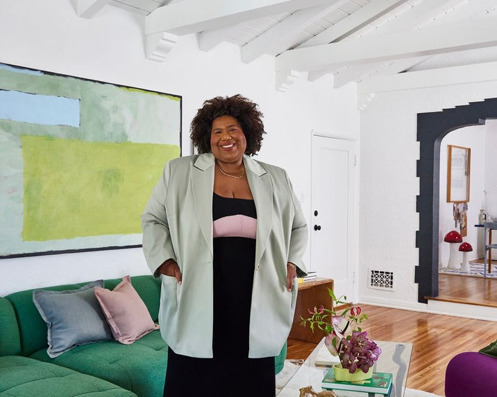 A smiling woman with curly hair stands in a stylish living room. She wears a light-colored blazer over a black outfit. Behind her are a green couch with pillows, a coffee table with plants, and abstract wall art.