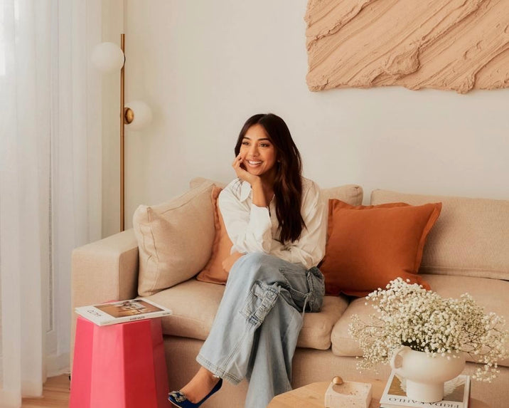 A woman with long brown hair sits smiling on a beige sofa with orange pillows in a cozy living room. She wears a white shirt and jeans. A pink side table, books, and a vase of white flowers are nearby. Abstract art hangs on the wall.