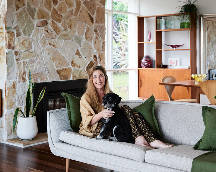 A woman with long hair sits on a light gray sofa, smiling and holding a black and white dog. The room features a stone fireplace, wooden floors, and mid-century modern furniture with large windows in the background.