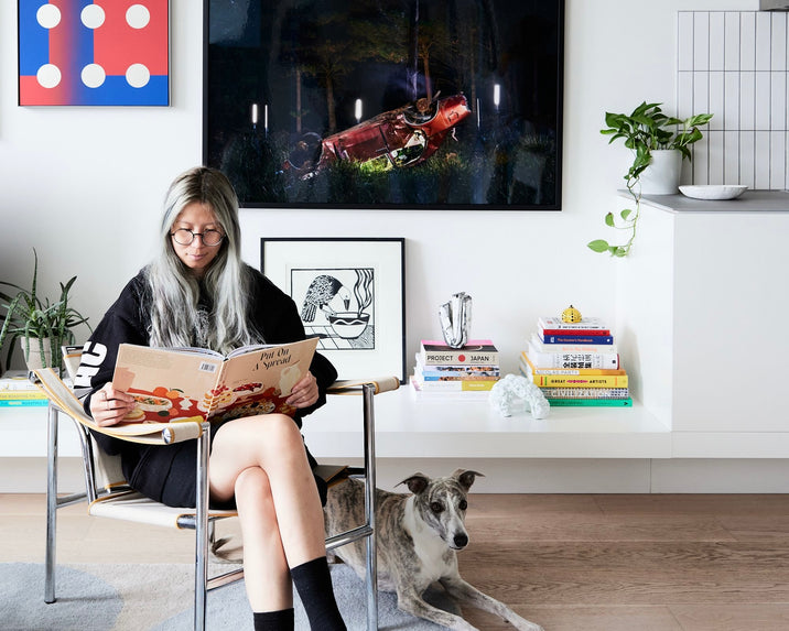 A woman with long gray hair sits in a modern chair reading a cookbook. A dog lies beside her on the floor. Behind them are books, plants, and colorful artwork on a white shelf and wall in a bright, stylish living room.