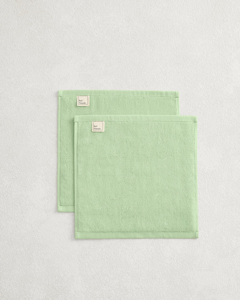The Bed Threads Apple 100% Cotton Terry Face Washers (Set of Two) are neatly folded, rectangular, light green with small beige labels, crafted from absorbent terry that's gentle on skin and placed on a light textured surface.
