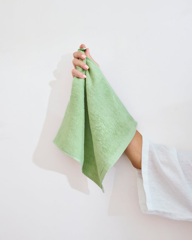 A hand with painted nails holds up a light green Bed Threads Apple 100% Cotton Terry Face Washer (Set of Two) against a white background, showing its gentle touch on skin. The person wears a long-sleeved white top.