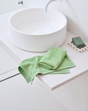 A modern white bathroom sink sits on a white countertop. Next to it are Bed Threads’ Apple 100% Cotton Terry Face Washers (Set of Two) and a matching green soap bar in a white dish, offering gentle care for your skin. - view