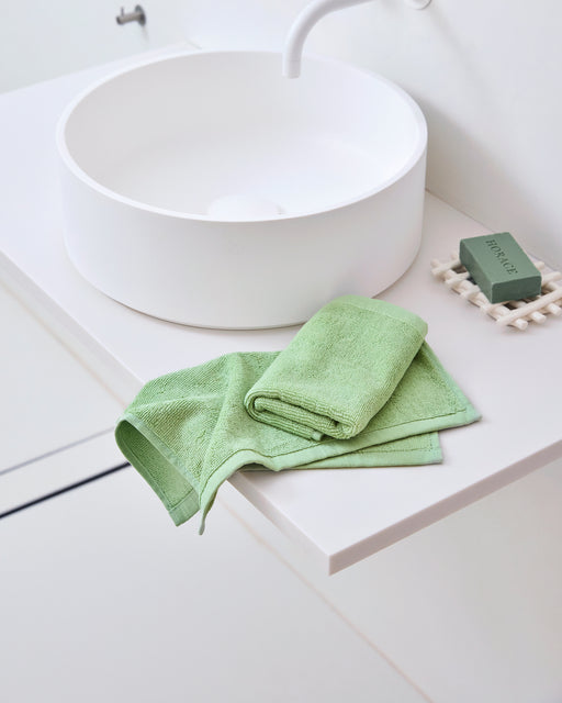 A modern white bathroom sink sits on a white countertop. Next to it are Bed Threads’ Apple 100% Cotton Terry Face Washers (Set of Two) and a matching green soap bar in a white dish, offering gentle care for your skin.