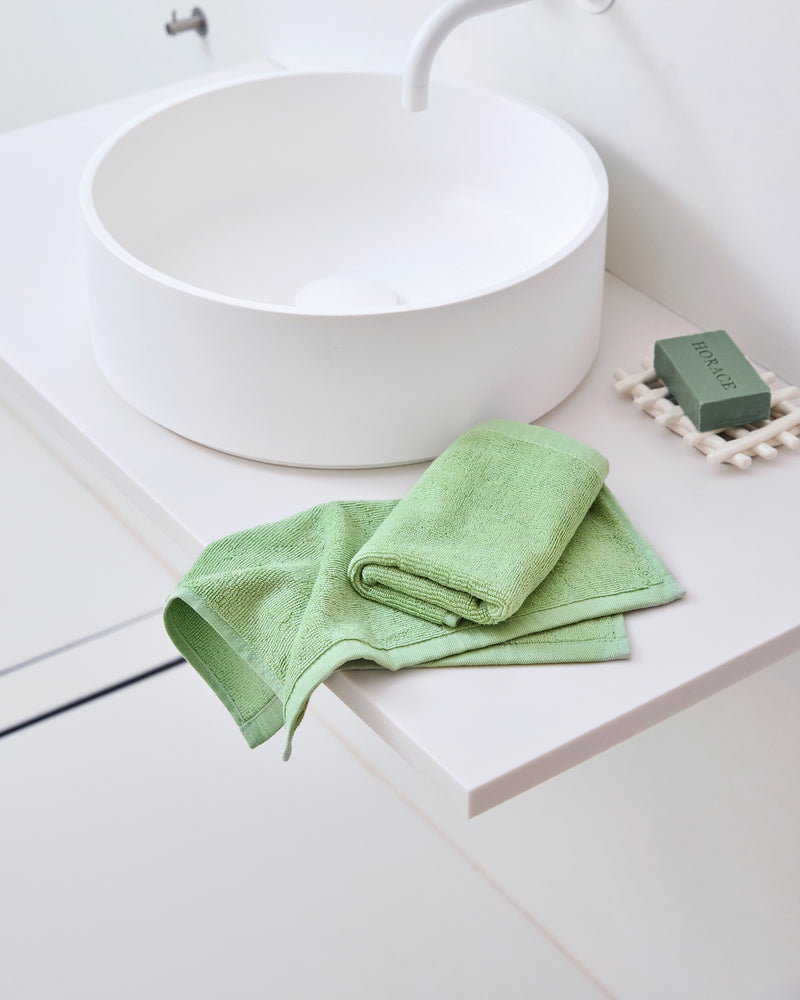 A modern white bathroom sink sits on a white countertop. Next to it are Bed Threads’ Apple 100% Cotton Terry Face Washers (Set of Two) and a matching green soap bar in a white dish, offering gentle care for your skin.
